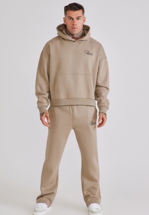 Hombre con tatuajes lleva sudadera con capucha beige y pantalones deportivos a juego con un pequeño logo negro, de pie frente a un fondo claro y liso.
