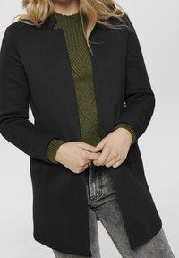 Manteau noir au design minimaliste, avec une coupe droite et sans attaches visibles, superposé à un pull vert texturé.