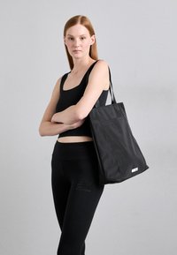 Geantă tote neagră cu o suprafață texturată, design structurat și mânere pentru umăr. Include un buzunar lateral mic și un accent cu etichetă albă.