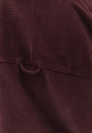 Bordeaux ribfluwel stof met verticale ribbels. Bevat een klein lusdetail aan de bovenkant nabij een geplooide naad.