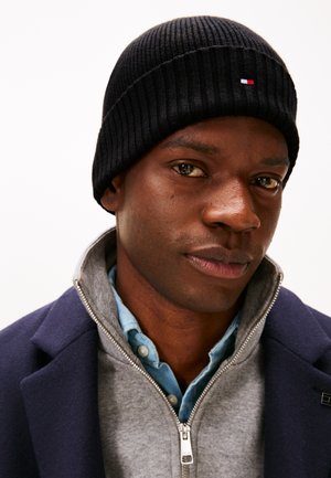 Homme portant un bonnet en maille noir, un manteau bleu marine, un pull gris zippé et une chemise bleue à boutons, faisant face à l'avant avec une expression neutre.