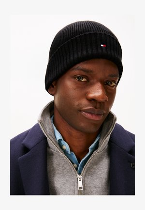 Homme portant un bonnet en maille noir, un manteau bleu marine, un pull gris zippé et une chemise bleue à boutons, faisant face à l'avant avec une expression neutre.