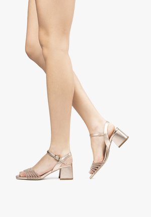 Sandales à talon bloc en rose doré métallique. Présentent un design de bride tressée, une boucle réglable à la cheville et un bout ouvert avec une texture lisse.