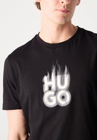 Černé bavlněné tričko s velkým bílým grafickým potiskem s textem "HUGO" ve stylu plamenů, kulatý výstřih, krátké rukávy.