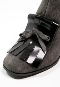 Gros plan sur une chaussure en daim gris avec un volant en cuir noir brillant et un nœud assorti noué à l'avant, au niveau des orteils.
