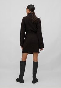 Vestido de malha castanho escuro com colarinho alto, mangas compridas e faixa na cintura. Comprimento até aos joelhos com punhos canelados e uma textura suave. Botas altas pretas até ao joelho.