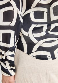 Poignet de chemise à manches longues à motif géométrique noir et blanc avec deux boutons noirs, portée avec un pantalon beige.
