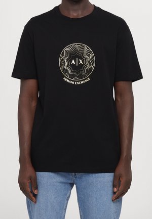 Mies pukeutuneena mustaan Armani Exchange T-paitaan, jossa on kullanvärinen pilkullinen pyöreä logo, ja vaaleansinisiin farkkuihin, seisoo yksivärisen taustan edessä.