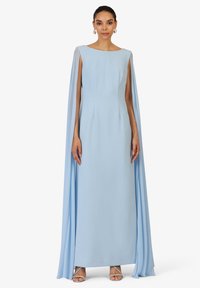 Robe longue bleu clair avec un décolleté large et des manches en forme de cape, réalisée en tissu lisse avec des coutures subtiles, associée à des chaussures à lanières.