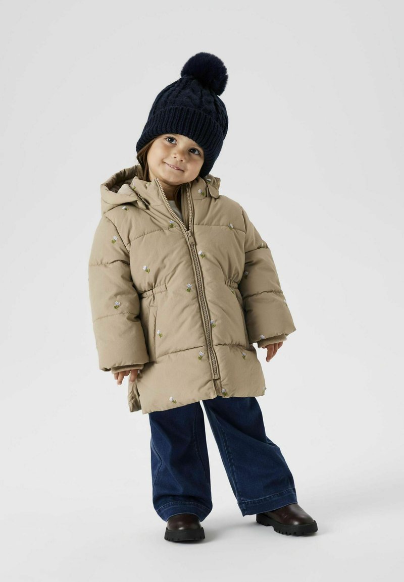 Manteau matelassé beige avec fermeture éclair et capuche, orné de broderies florales. Associé à un pantalon en denim foncé à jambes évasées et un bonnet en tricot bleu marine avec un pompon.