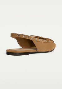 Hnědé semišové ploché muly se slingback designem, kovovými očky podél horního okraje a čtveratou špičkou na bílém pozadí.