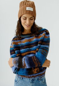 Pull en tricot avec un motif rayé multicolore en bleu, marron et noir, avec un col rond et des manches longues. Associé à un bonnet côtelé marron.