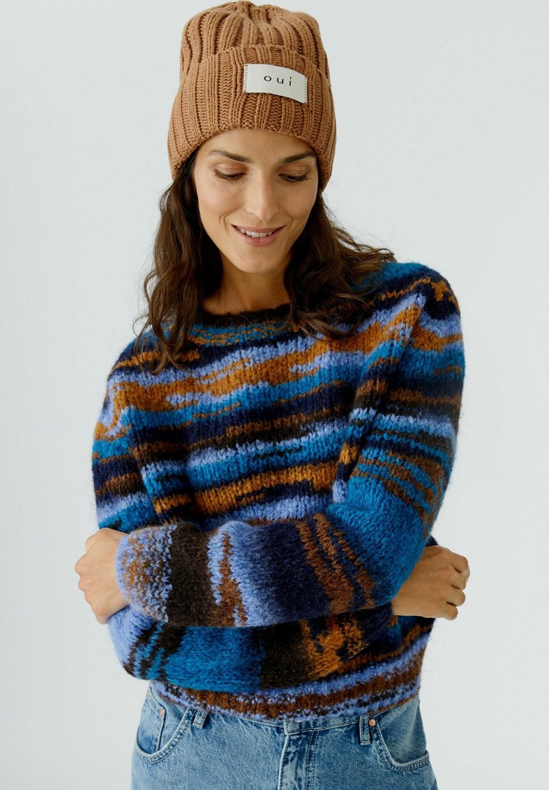 Pull en tricot avec un motif rayé multicolore en bleu, marron et noir, avec un col rond et des manches longues. Associé à un bonnet côtelé marron.