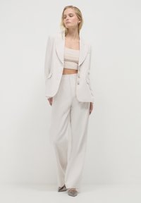 Frau in einem cremefarbenen Blazer, dazu passenden weit geschnittenen Hosen, einem beigen Crop-Top und spitzen metallisch silbernen Heels vor einem schlichten Hintergrund.