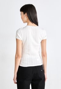 Hvit ribbestrikket skjorte med korte ermer, har blonder på halsen og ermene. Brukt med svarte høyt midjepants, som viser en tettsittende design.
