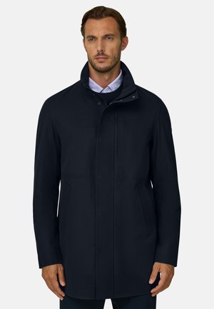 Homme portant un manteau noir mi-long à col montant sur une chemise bleu clair et un pull noir, sur un fond clair uni.