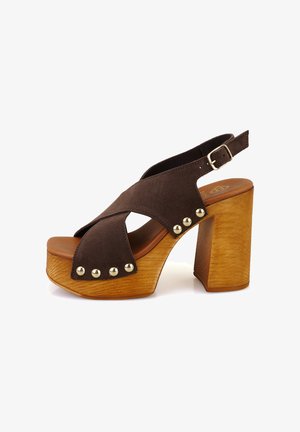Braune Wildleder-Plateausandalen mit überkreuzten Riemen, einem Holzblockabsatz und goldfarbenen Nieten, die die Vorderseite akzentuieren. Schnallenverschluss an der Ferse.