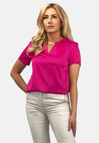 Różowa bluzka z krótkim rękawem z dekoltem w kształcie łezki i fałdami przy dekolcie, zestawiona z jasnymi, dopasowanymi spodniami. Gładka struktura materiału.