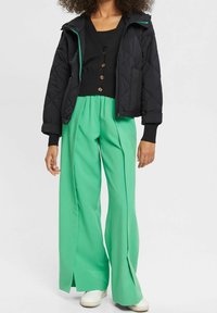 Giacca nera trapuntata con dettagli teal, indossata sopra un top nero a maniche lunghe e pantaloni verde a gamba larga con fessure frontali. Sneakers bianche.