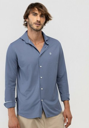 Hombre con cabello castaño claro que lleva una camisa azul de manga larga abotonada y pantalones beige sobre un fondo claro y liso.