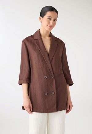 Donna con capelli scuri pettinati all'indietro, indossa un blazer marrone doppiopetto largo e pantaloni bianchi su uno sfondo semplice.