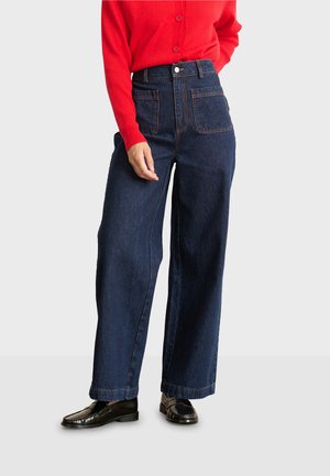 Donkerblauwe wijde denim jeans met een hoge taille, zichtbare stiksels en twee achterzakken. Gecombineerd met een rode geribde cardigan.