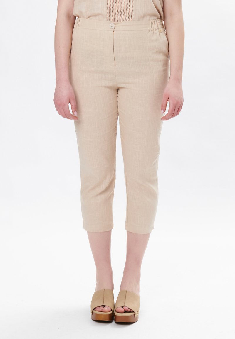 Pantaloni capri beige realizzati in un tessuto testurizzato, con una vita elastica e un unico bottone. Abbinati a scarpe con plateau e punta aperta.