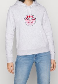 Felpa grigia chiara con tasca frontale, cappuccio con cordino e logo grafico in rosso e nero. Tessuto morbido, polsini e orlo a coste.