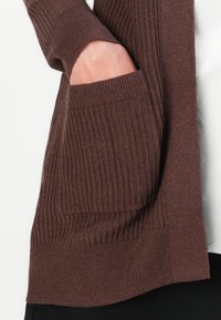 Hand inserted into the ribbed knit pocket of a brown cardigan worn over a white shirt and black pants.