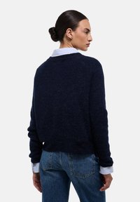 Pull en maille bleu marine avec une coupe décontractée, des poignets côtelés et un col rond, porté sur une chemise blanche à col, associé à un jeans en denim.