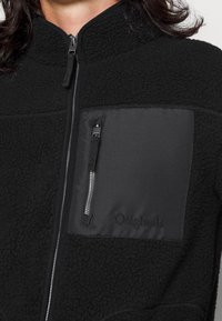 Veste polaire noire avec col montant, fermeture éclair et poche poitrine gauche. Tissu texturé avec des accents lisses ; logo sur la poche.