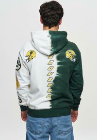 Huppari, jossa on sidontakuvio, vihreä-valkoisissa väreissä, NFL-logot hihoissa ja keltainen teksti "RECOVERED".