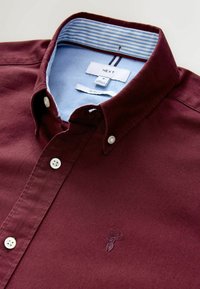 Camisa burdeos de botones con un diseño estilizado, que presenta un cuello interior a rayas azul claro y un pequeño logotipo bordado.