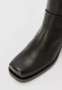 Botín negro de cuero con punta cuadrada, forma sencilla y textura suave con un ligero brillo. Acentuado con una banda estrecha alrededor del eje.