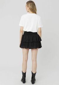 Jupe noire à volants avec un ourlet en dentelle, dotée de détails en œillets. Associée à un t-shirt blanc. La mannequin porte des bottines noires.