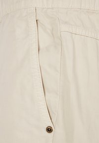 Beige stof med en tekstureret vævning, der har en sidelomme og en dekorativ metaltrykknap. Designet er enkelt med rene linjer.