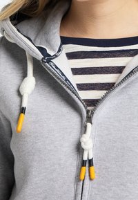 Felpa con zip grigia dal finish testurizzato. Presenta lacci bianchi con accenti gialli e neri, indossata sopra una maglietta a righe.