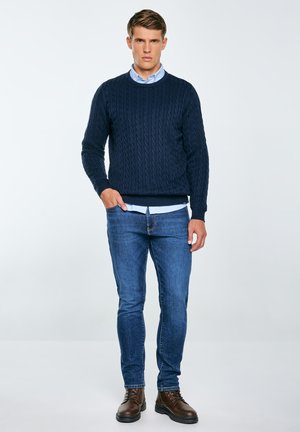 Granatowy sweter w splot kablowy z okrągłym dekoltem, nałożony na jasnoniebieską koszulę, w połączeniu z niebieskimi dżinsami i brązowymi butami na sznurówki.