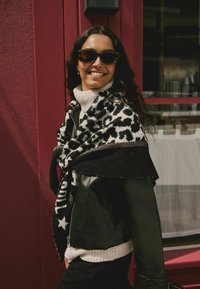 Sort læderjakke over en cremefarvet rullekrave, draperet med et sort-hvidt leopardprint sjal med metalaccenter. Baggrunden er pink.