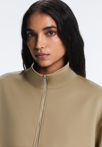 Femme aux longs cheveux foncés portant une veste beige zippée à col montant, regardant droit devant sur un fond clair uni.