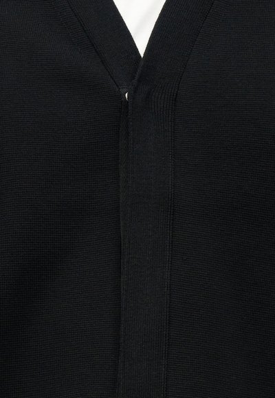 Μαύρο πλεκτό κοντόζακα με λαιμόκοψη σε σχήμα V φορεμένο πάνω από λευκό πουκάμισο, με λεπτή ριμπ υφή και μερικά κουμπιά κλειστά κοντά στο λαιμό.