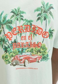 Baltas T-krekls ar krāsainu grafiku, kurā attēlots sarkans klasiskā automašīna, palmas un teksts "PERDIDO en el PARAÍSO" treknā sarkanā un melnā krāsā.