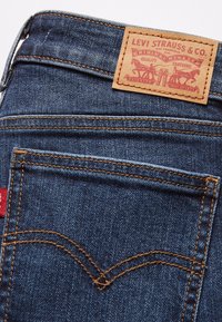 Jeans de mezclilla con un lavado azul oscuro. Presentan una etiqueta de cuero marrón con texto rojo, costuras amarillas y dos bolsillos traseros de diseño clásico.