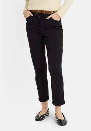 Mujer con pantalones negros remangados, jersey beige, cinturón marrón y zapatos planos negros con tachuelas, con las manos en los bolsillos.