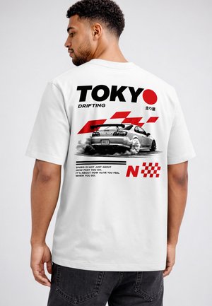 Man wearing white T-shirt with graphic of drifting car, red racing stripes, and text "TOKYO DRIFTING" on the back against plain background.