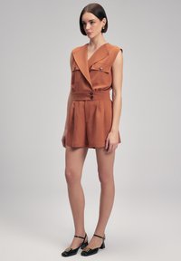 Rostorange ärmelloser Jumpsuit mit Schalkragen, Fronttaschen und einem Stoffgürtel; kombiniert mit schwarzen Schuhen mit Absatz und goldenen Akzenten.