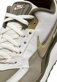 Zapatilla Nike blanca con un diseño texturizado y acentos en beige y verde olivo. Incluye una lengüeta acolchada con el logo y cordones planos.