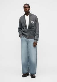 Άνδρας που φορά γκρι πουλόβερ με φερμουάρ και ανάγλυφη υφή, με μαύρο κείμενο, λευκό πουκάμισο, φαρδιά ανοιχτό μπλε τζιν και σκούρα παπούτσια, στέκεται μπροστά σε μονόχρωμο φόντο.