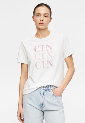 Kobieta w białej koszulce z napisem „CIN” powtórzonym trzy razy różowymi literami, lekko wpuszczoną w jasnoniebieskie jeansy z ręką w kieszeni.