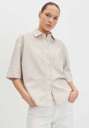 Frau mit zurückgebundenem Haar, trägt ein hellbeiges, kurzärmeliges, perforiertes Hemd mit Knöpfen und weiße Hosen, Hände in den Taschen, neutraler Hintergrund.